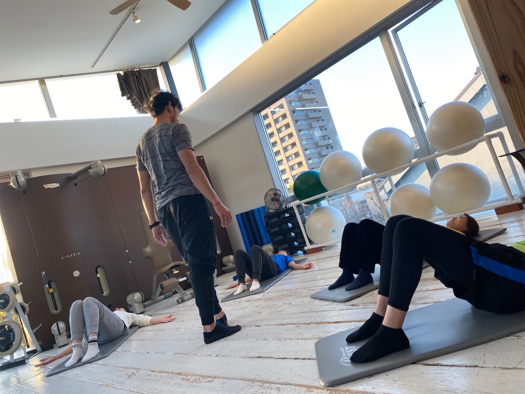 スタジオでグループレッスンを指導するトレーナーと、マットに寝て動作を行う会員